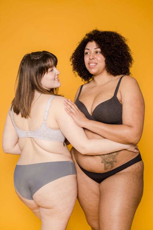 Free Positive plus size African American woman and Asian overweight female in underwear hugging while standing on yellow background in studio Stock Photo