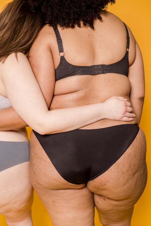 Back view of multiracial anonymous plus size female models wearing lingerie and hugging while standing on yellow background in light studio
