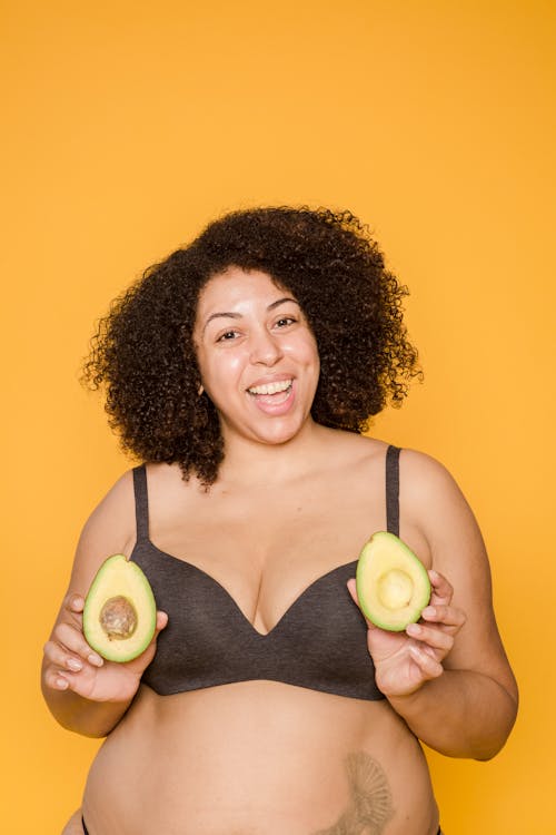 Cheerful plus size African American female wearing lingerie smiling and looking at camera while standing on yellow background with halves of ripe avocado