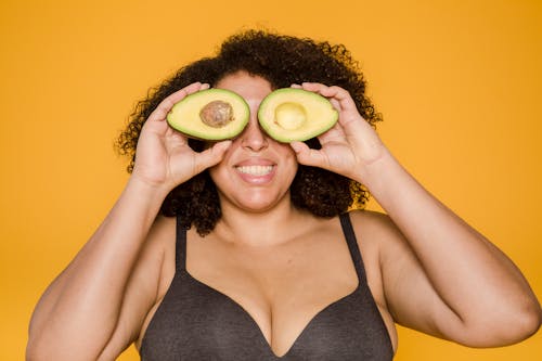 Cheerful overweight ethic model with cut avocado on yellow background