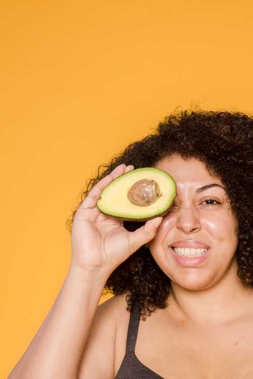 Free Cheerful plump African American female with curly hair covering eye with cut avocado while looking at camera Stock Photo