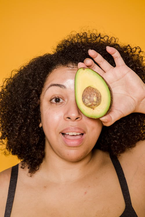 Free Amazed African American female with curly hair covering eye with cut avocado while looking at camera on yellow background Stock Photo