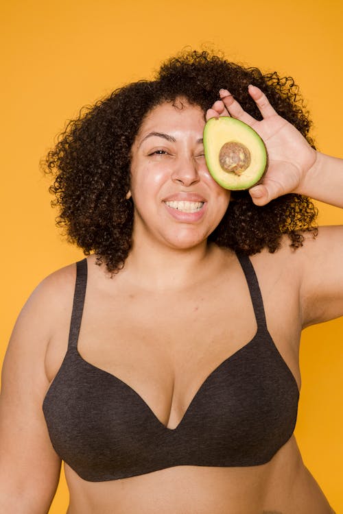 Cheerful overweight ethnic model in bra with cut avocado
