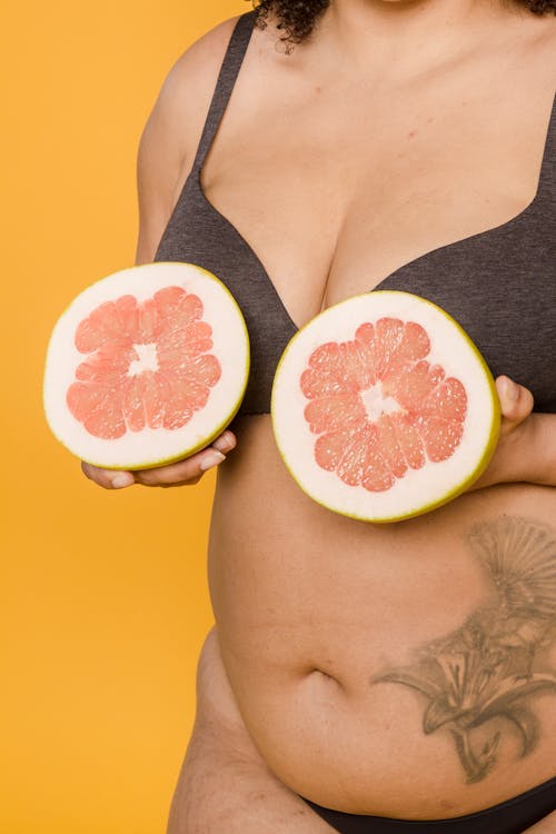 Crop anonymous plus size woman in underwear with tattoo and juicy grapefruit halves on yellow background