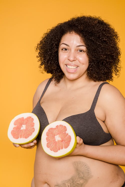 Cheerful plus size African American female in brassiere with tattoo and fresh grapefruit halves looking at camera on yellow background