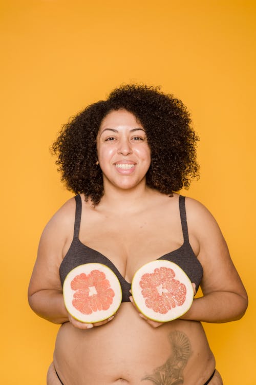 Cheerful plump African American woman in lingerie with tattoo and juicy grapefruit halves looking at camera on yellow background