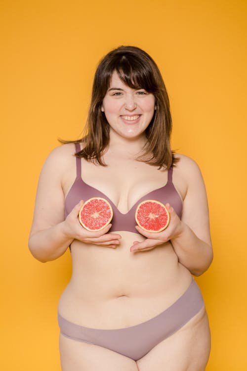 Cheerful plus size Asian woman in lingerie with juicy grapefruit halves looking at camera on yellow background