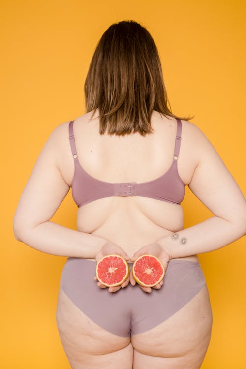 Back view of anonymous plump woman in underwear with juicy grapefruit halves and skin folds on yellow background