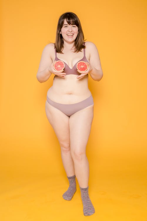 Cheerful plump ethnic female model in lingerie and socks with grapefruit halves looking at camera on yellow background