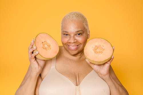 Cheerful plus size African American woman in brassiere with fresh melon halves looking at camera on yellow background