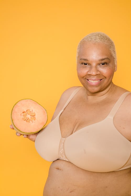 Cheerful mature plus size African American woman in bra with fresh melon half looking at camera on yellow background