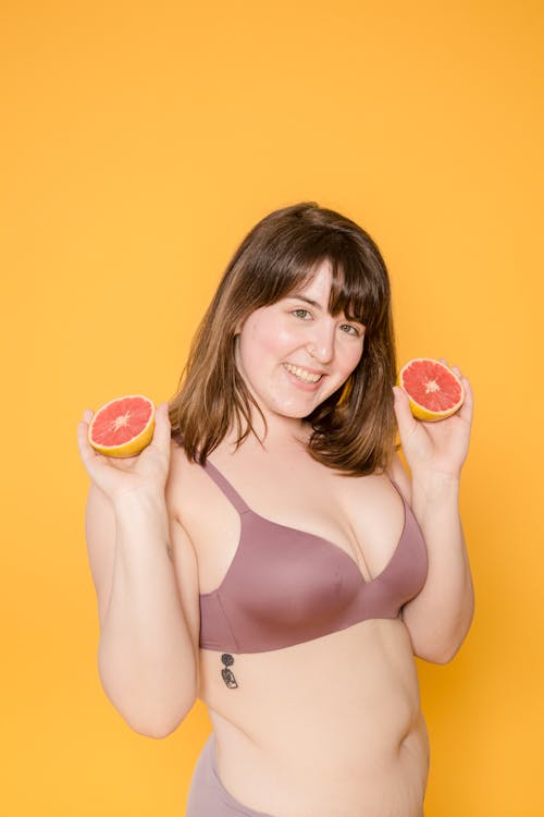 Cheerful overweight ethnic female in lingerie with cut fresh grapefruit and tattoo looking at camera on yellow background