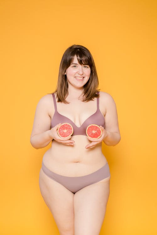 Cheerful plus size ethnic woman in bikini and brassiere with cut grapefruit looking at camera on yellow background