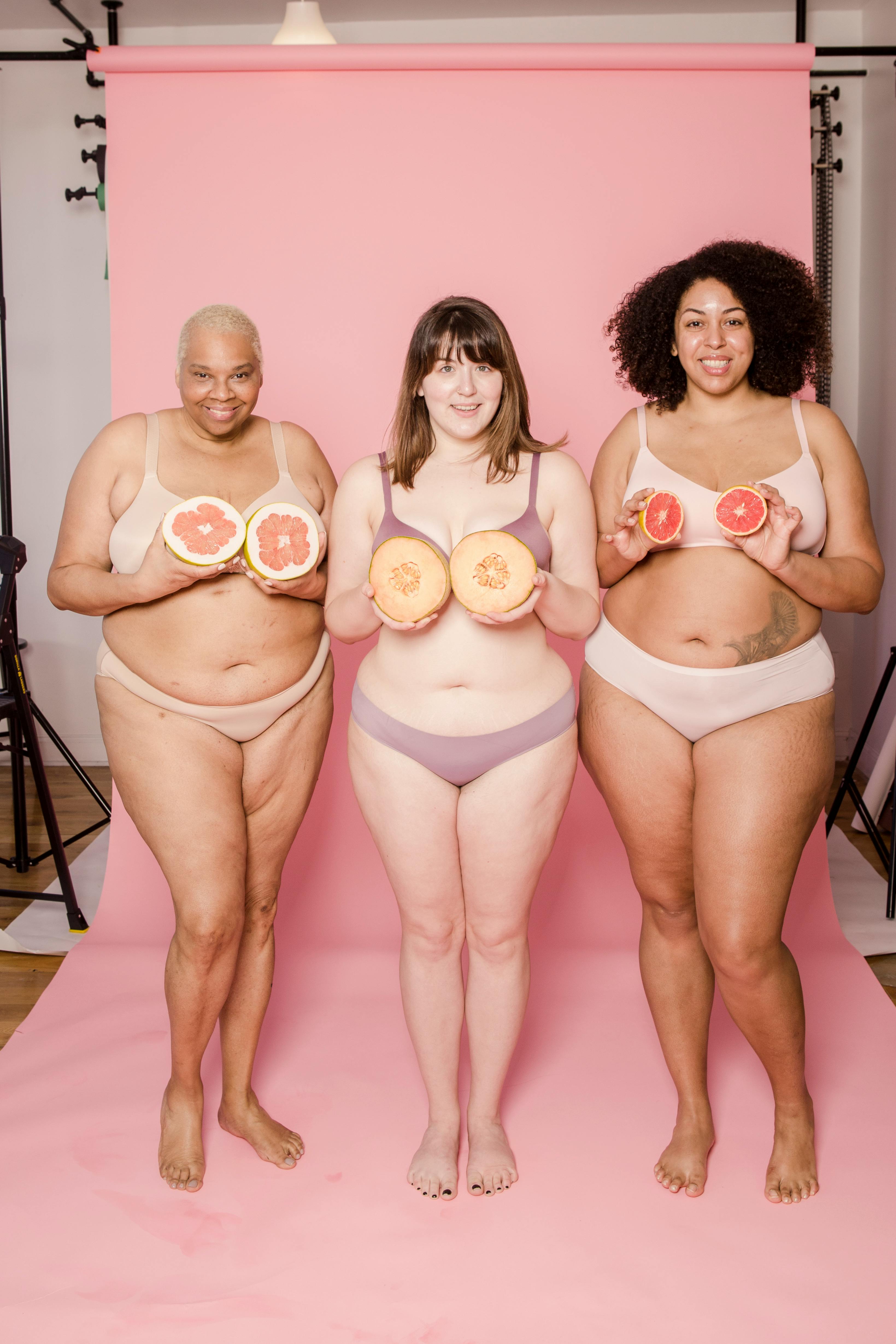 smiling plump multiracial girlfriends in underclothes with cut fruits indoors