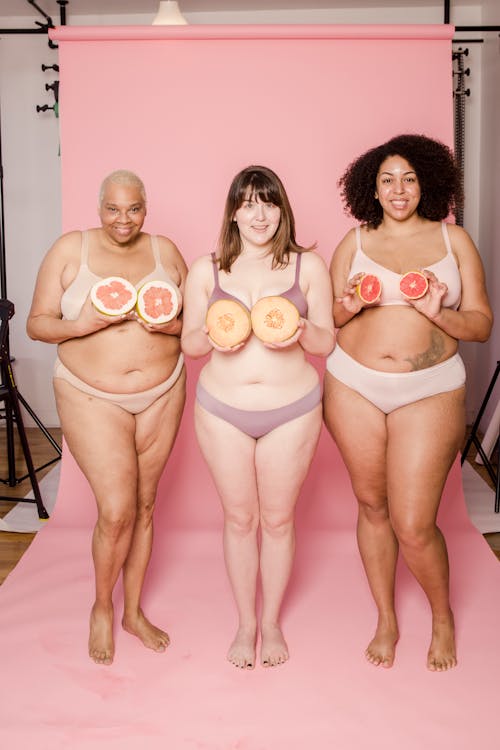 Cheerful plus size multiethnic models in lingerie with fresh fruit halves looking at camera in photo studio