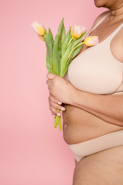 Side view of unrecognizable overweight African American female model in underwear with bouquet of fresh flowers on pink background in studio