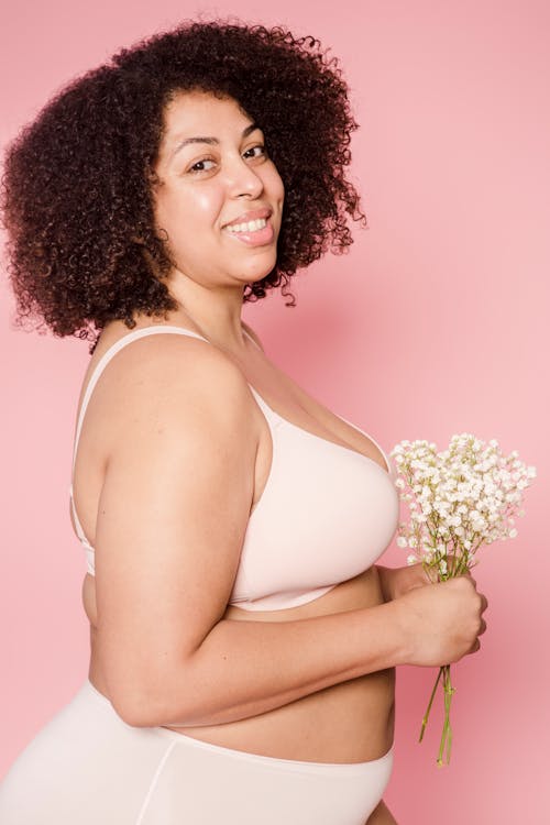 Cheerful plump African American female with black hair wearing lingerie standing on pink background with small flowers in hands while looking at camera