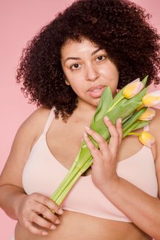 Plus size African American female model wearing beige lingerie looking at camera on pink background with fresh tulips in hands