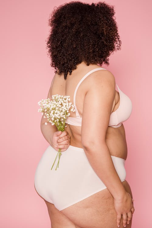 Side view of faceless plus size African American female in beige lingerie with flowers behind back standing on pink background in studio