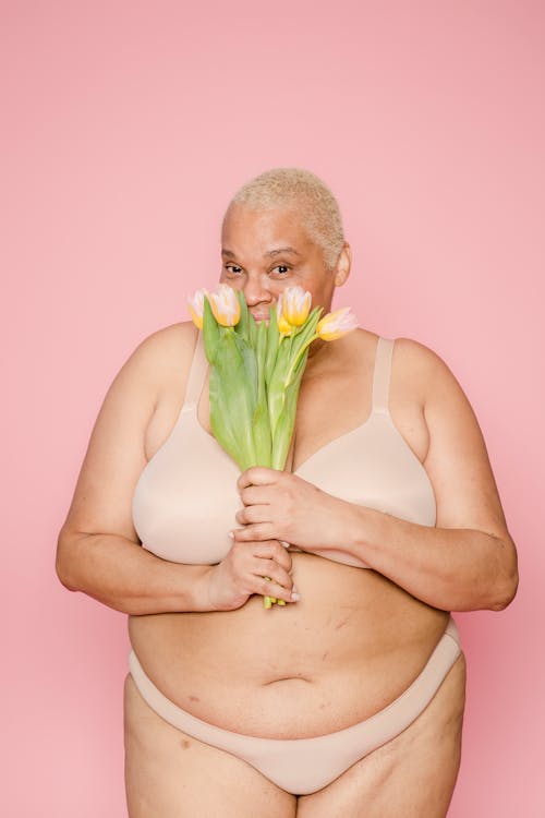 Cheerful overweight African American female model in lingerie covering half face with fresh tulips on pink background in light studio