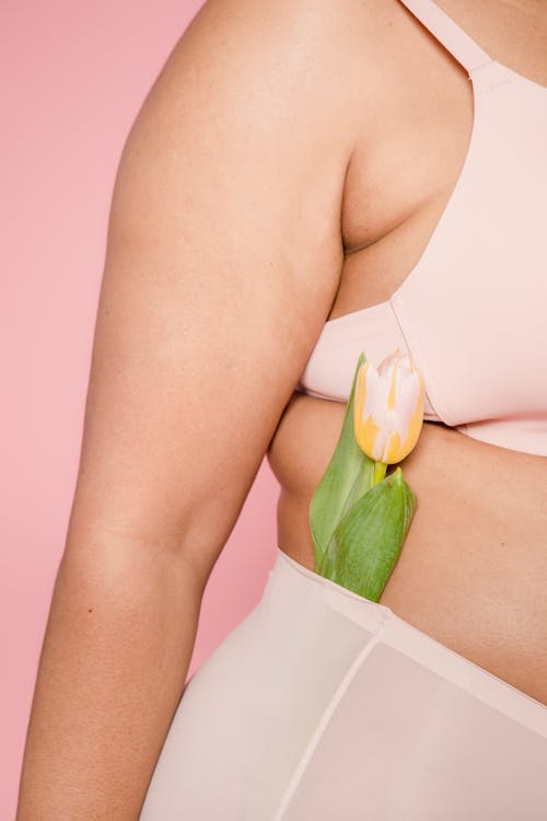 Unrecognizable plus size female model wearing beige underwear standing on pink background with fresh tulip in panties in light studio