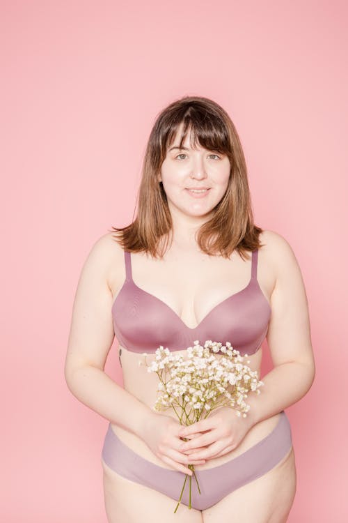 Positive plus size Asian female model in lingerie with small flowers in hands looking at camera on pink background in studio