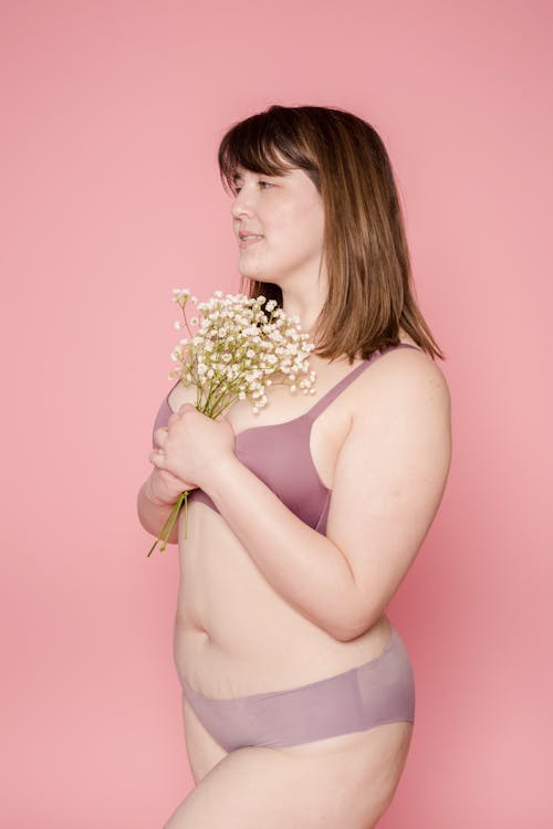 Free Side view of peaceful plump Asian female model in lingerie standing on pink background with flowers in hands in studio Stock Photo