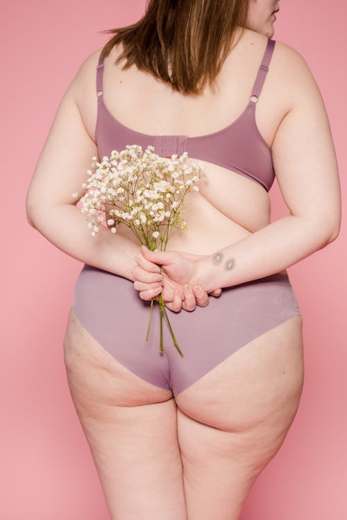 Back view of unrecognizable plump female in lingerie with bouquet of fresh small flowers in hands standing on pink background