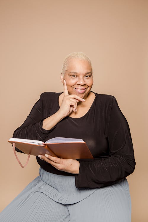 Happy overweight African American female with opened notebook in hand smiling and looking at camera on beige background in studio