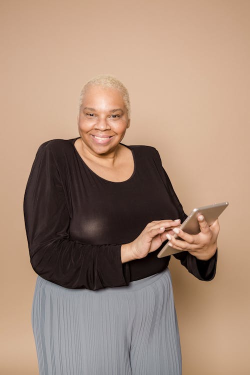Cheerful overweight African American female with short hair browsing tablet and looking at camera on beige background in light studio