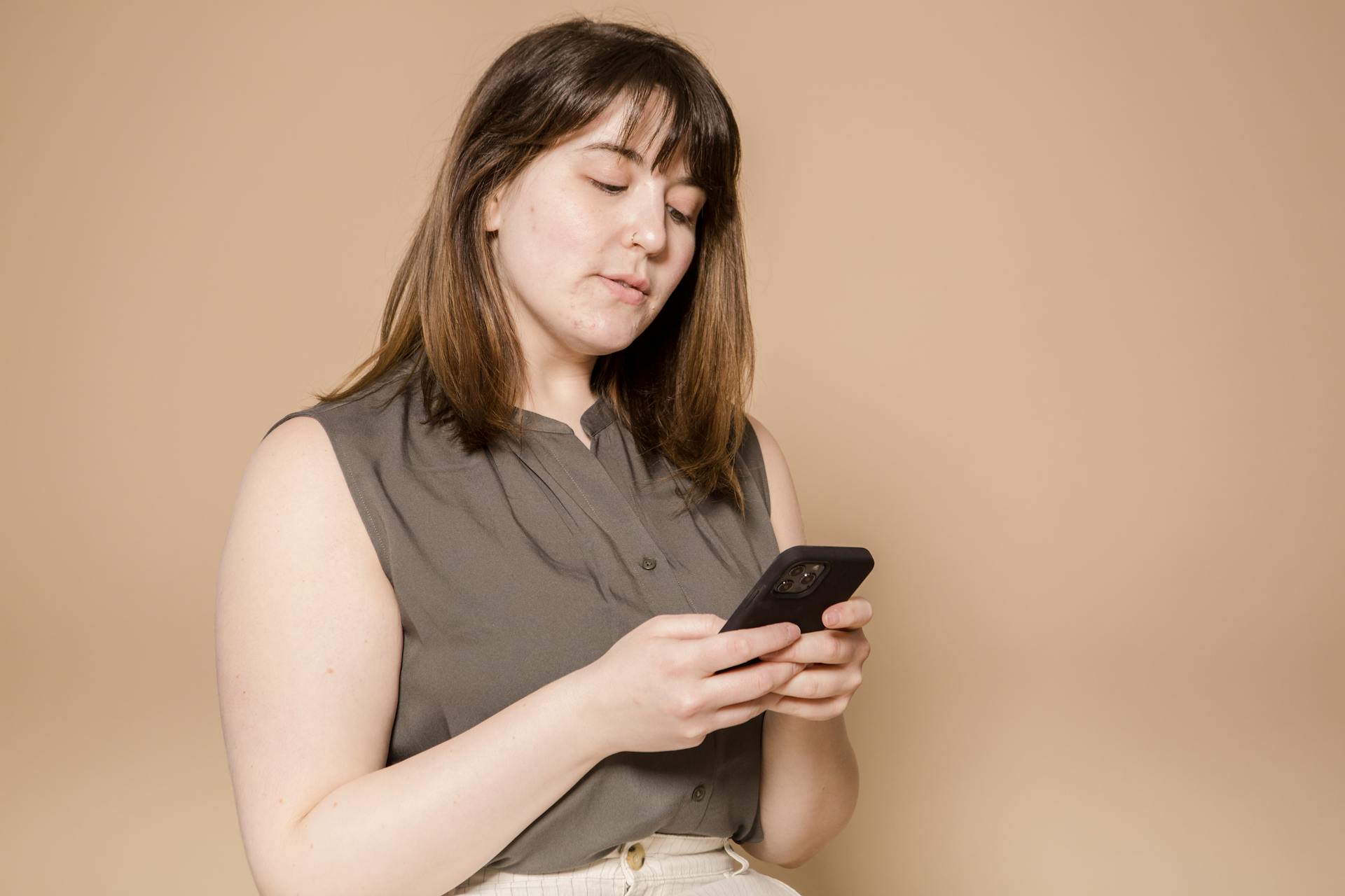 Focused plump Asian female in stylish wear text messaging on modern cellphone while sitting on beige background in light studio