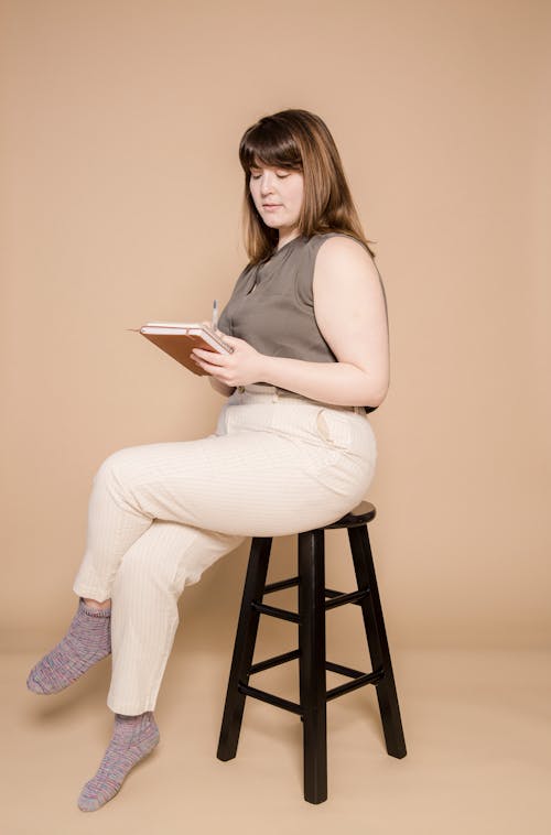 Full body of focused overweight Asian female taking notes in notepad while sitting on chair against beige background in studio