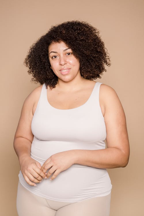 Optimistic plump African American female with curly hair wearing white t shirt looking at camera on beige background in studio