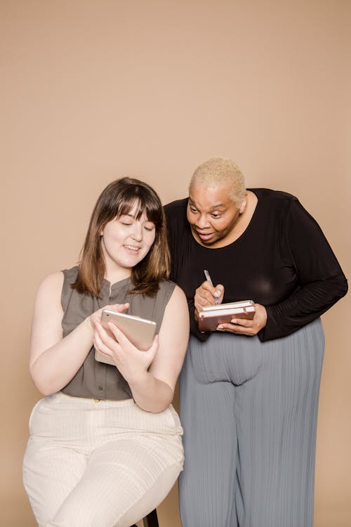 Positive Asian woman showing tablet to amazed plump African American female colleague with notebook while looking at screen on beige background in studio