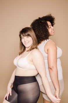Joyful plump Asian woman in lingerie looking at camera while standing back to back with overweight African American female on beige background