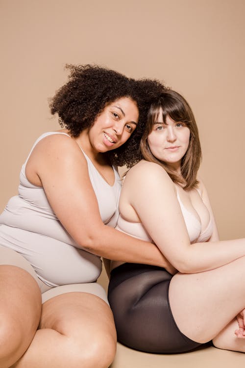 Cheerful plus size multiethnic female models wearing underwear embracing and looking at camera while sitting on beige background in studio