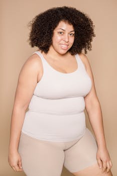 Delighted plump African American female with black hair wearing shorts smiling and looking at camera on beige background in studio