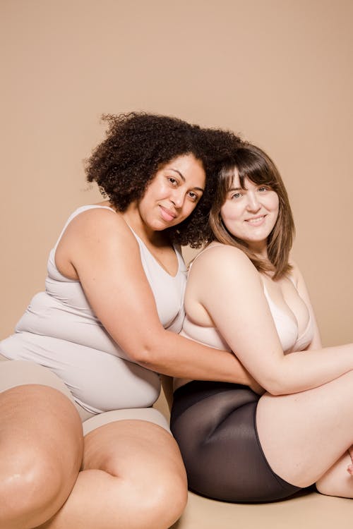 Overweight multiethnic female models in underwear smiling and looking at camera while embracing on floor against beige background in studio