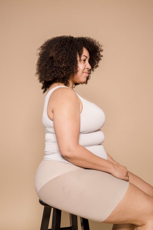 Side view of cheerful plus size African American female wearing shorts and t shirt sitting on chair against beige background