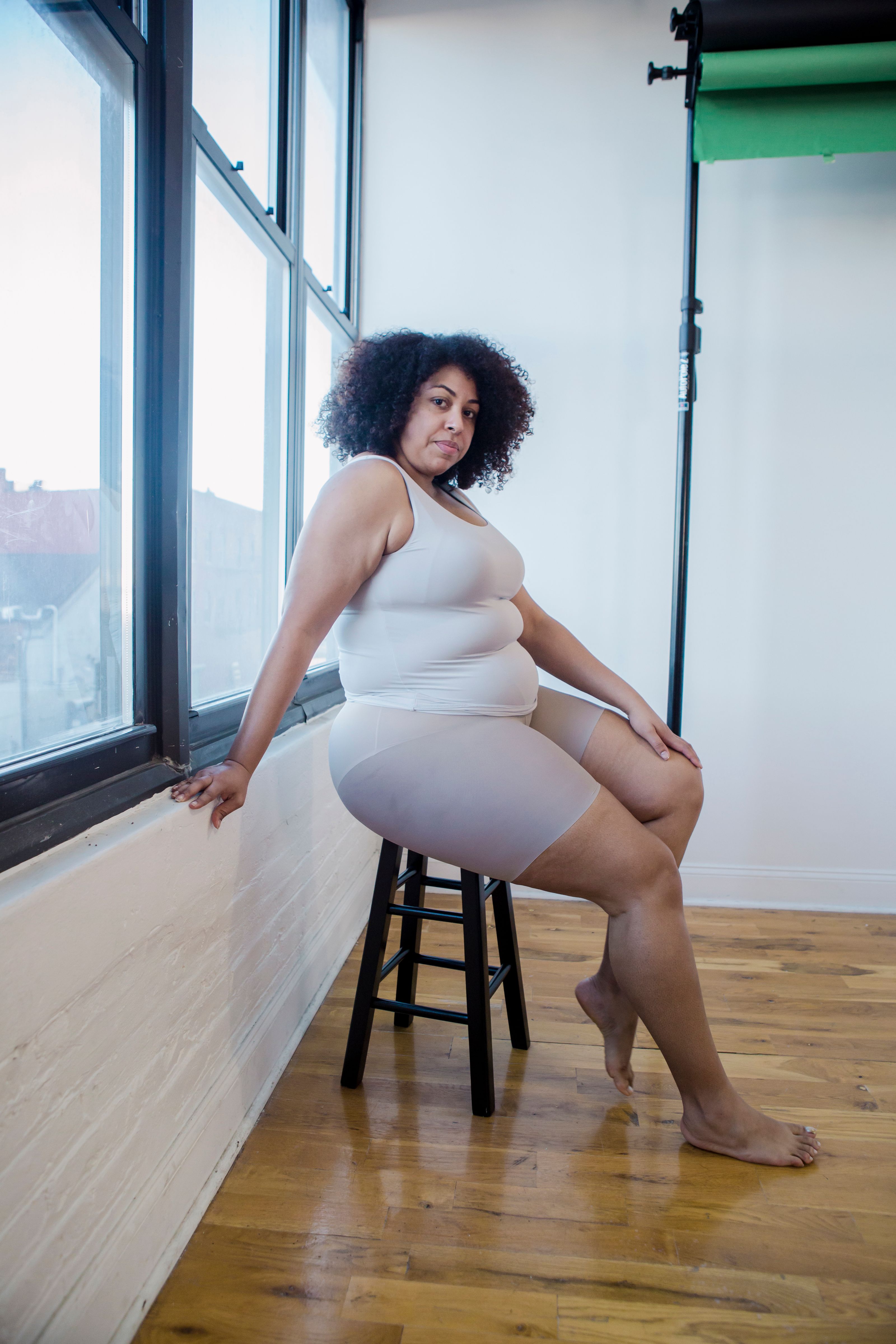 Mulher plus size com cabelo cacheado senta-se confiantemente em um banquinho. A luz natural entra por grandes janelas.