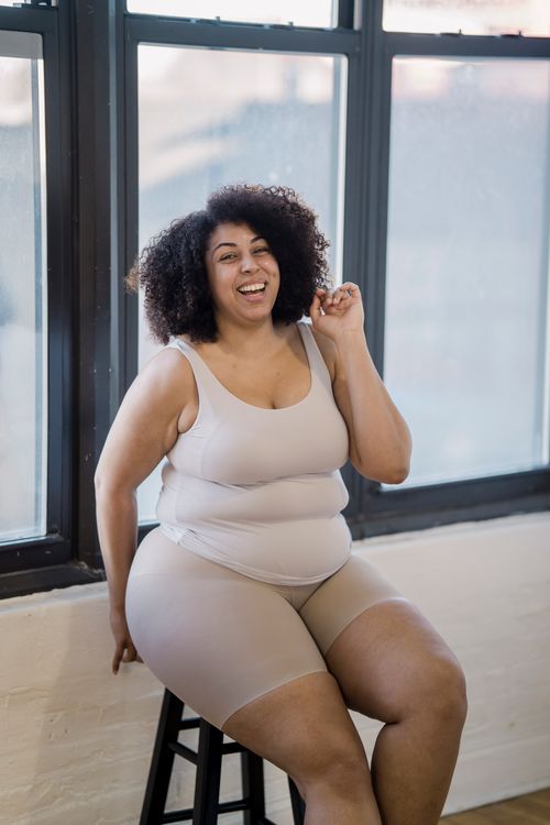 Free Cheerful plump African American woman in underwear touching curly hair while looking at camera and sitting on stool Stock Photo