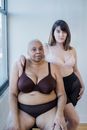 Asian woman embracing mature plus size African American girlfriend with scar in lingerie while looking at camera indoors