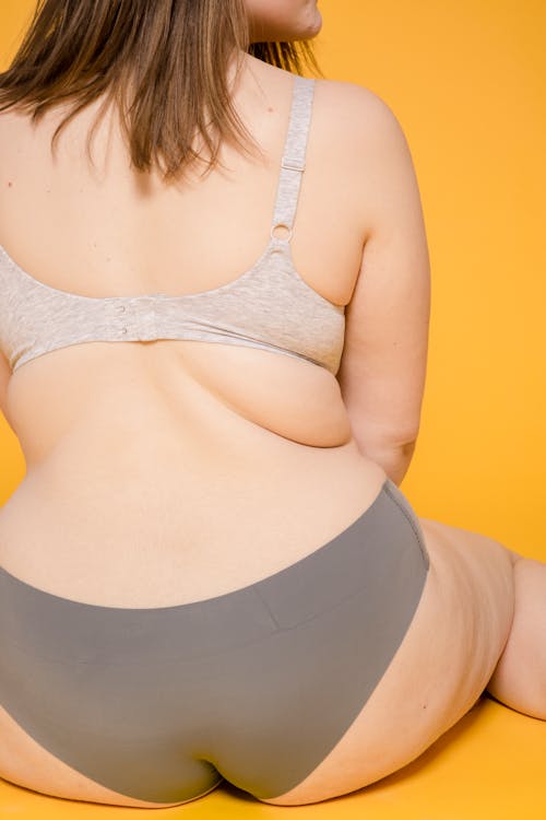 Free Back view of crop anonymous woman with skin fold in panties and brassiere sitting on yellow background Stock Photo