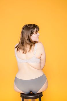 Back view of young plus size woman in panties and bra sitting on stool while looking away