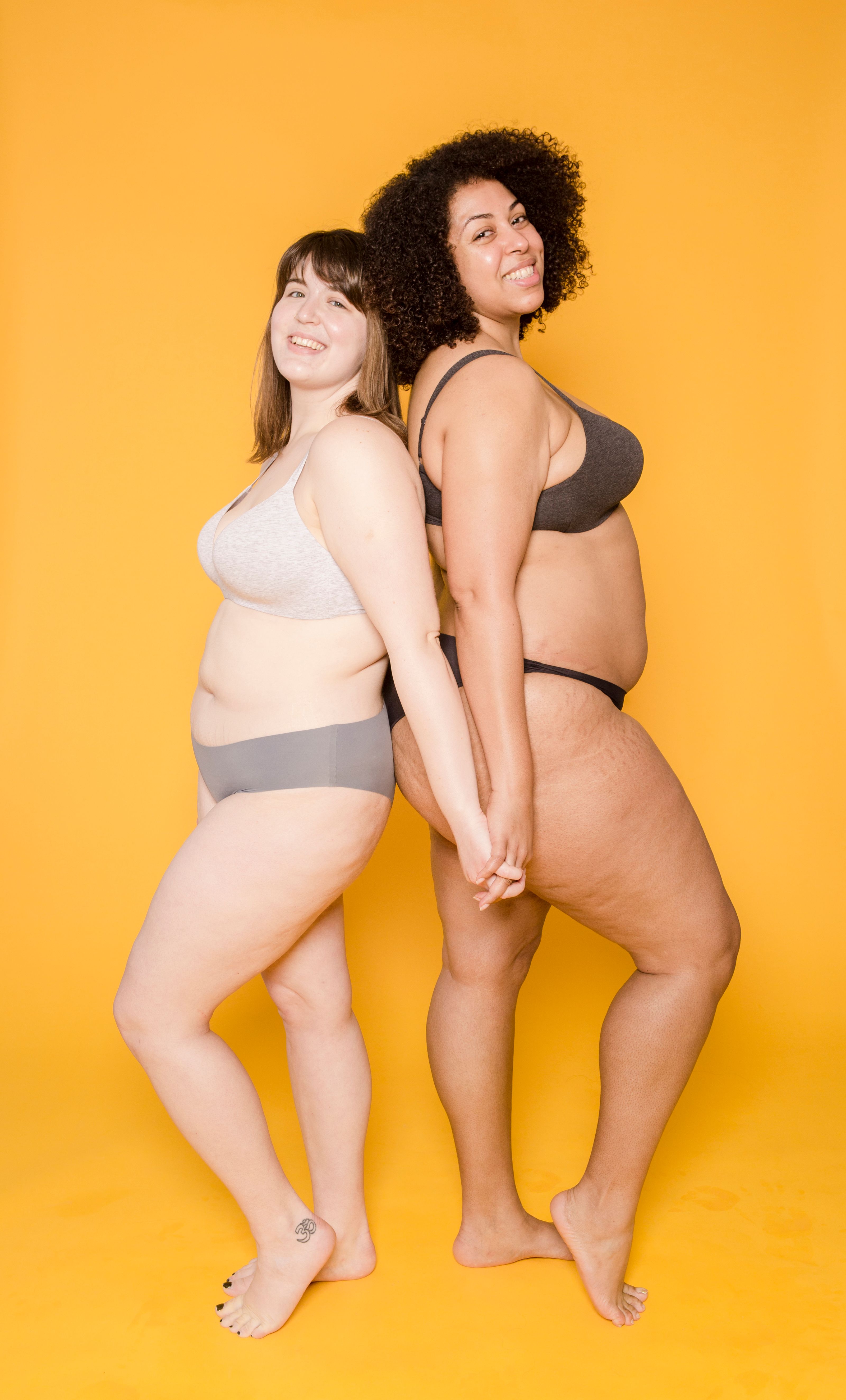Duas mulheres diversas em roupas íntimas sorrindo confiantemente contra um fundo amarelo brilhante.