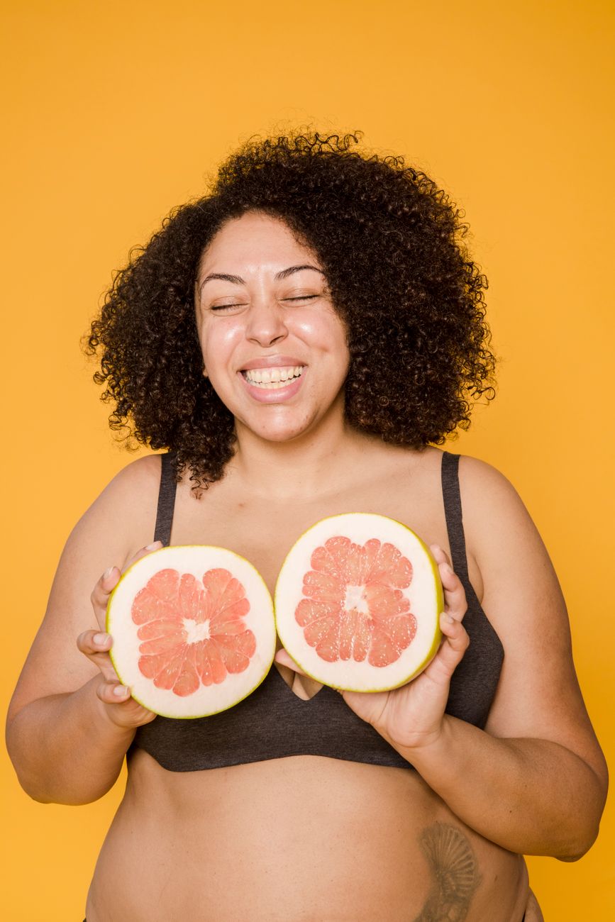 Smiling woman with curly hair holds sliced grapefruits against a vibrant yellow background.
