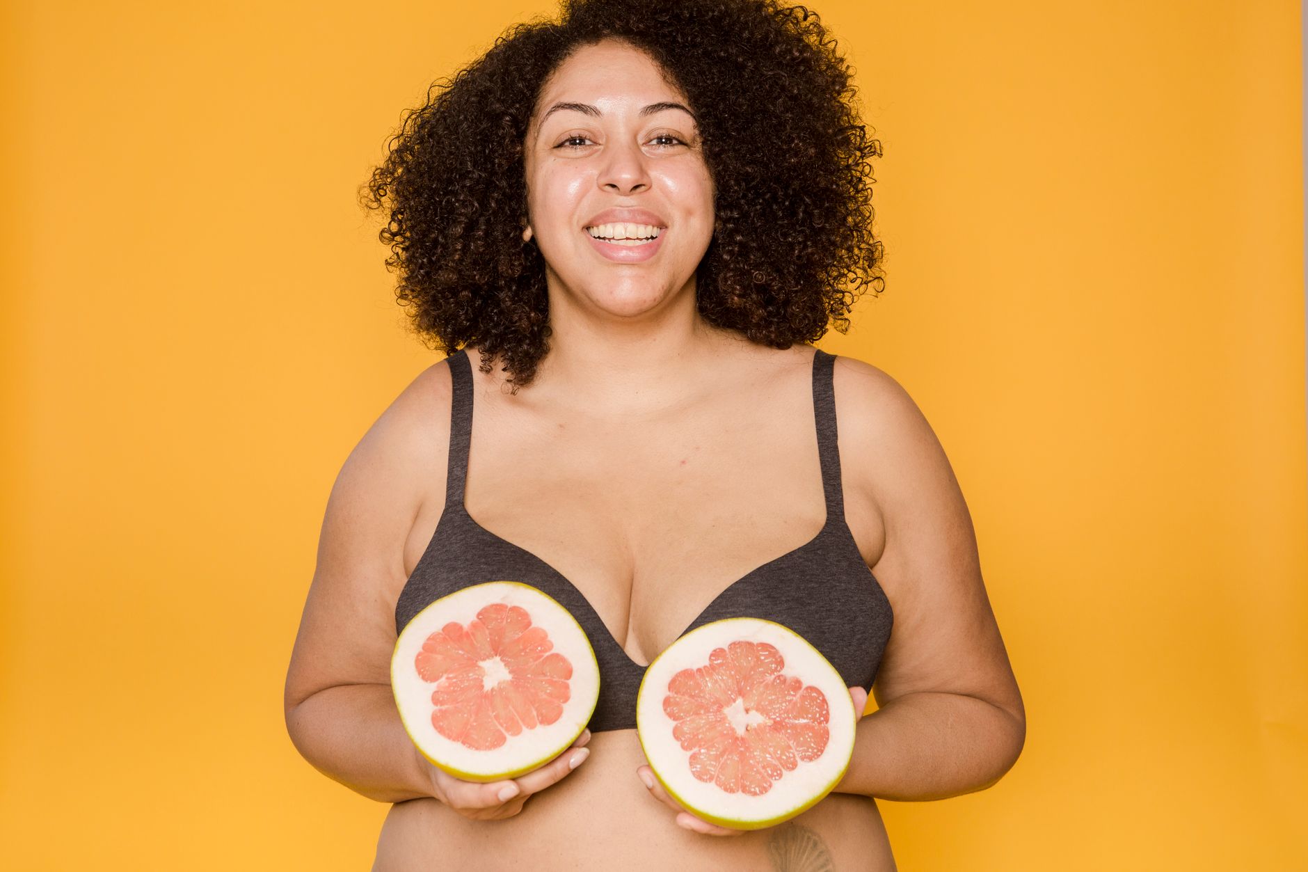 Confident smiling woman with curls holding grapefruit slices on a vibrant yellow background.