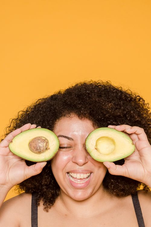 Free Cheerful woman with curly hair playfully holding avocado halves in front of eyes against a vibrant yellow backdrop. Stock Photo