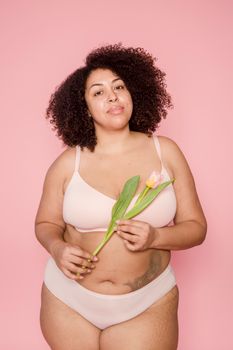 Empowered plus size woman with curly hair holding a tulip against a pink background.