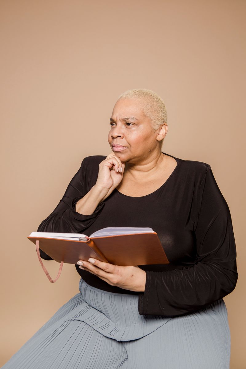 A midlife Black woman with short platinum-blonde hair holds an open journal in her lap, hand to chin, gazing thoughtfully off-frame against a warm tan studio backdrop.